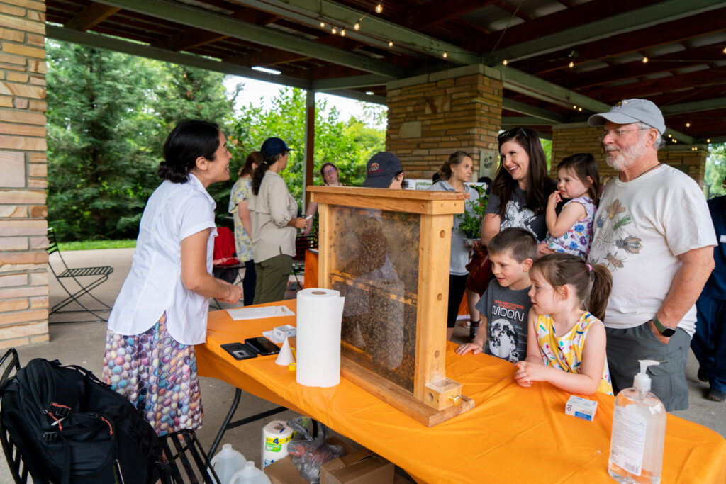 A woman talks to children and adults at a table with a beehive on it