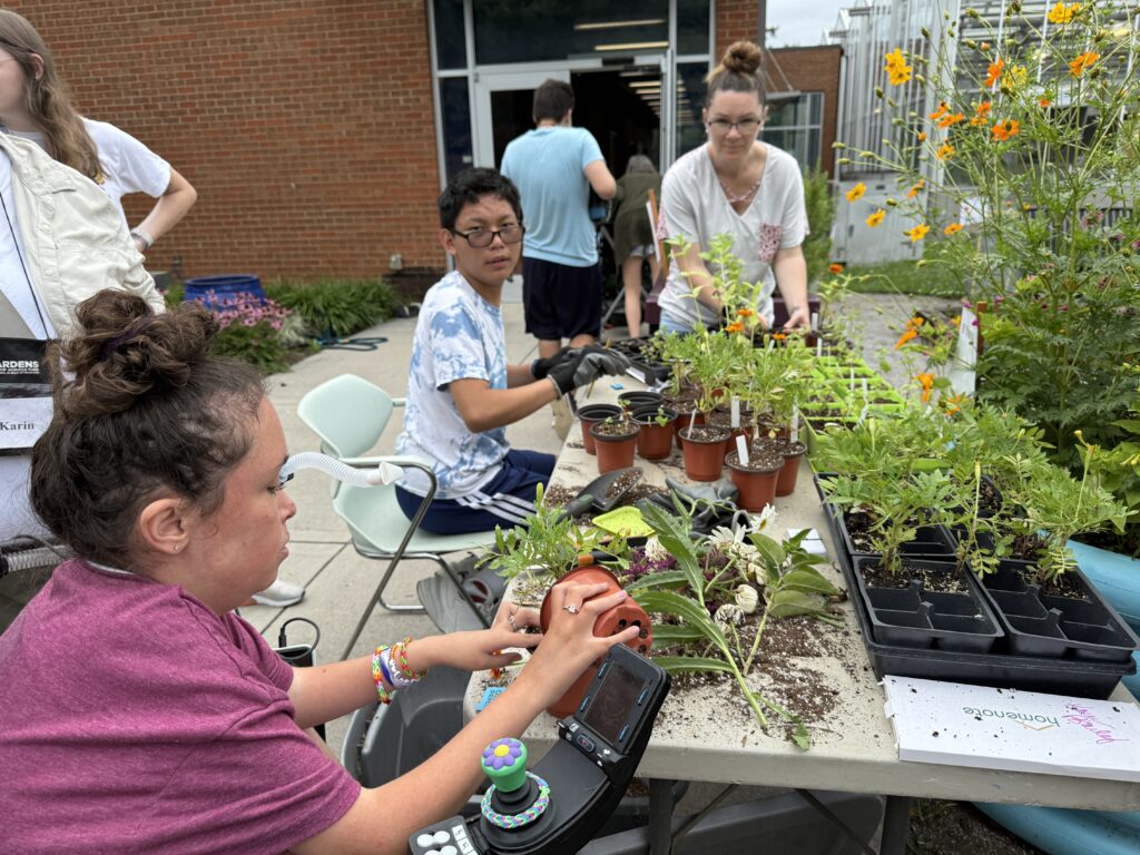 Image of teenagers and an adult transplanting various plants at a table that is outside.