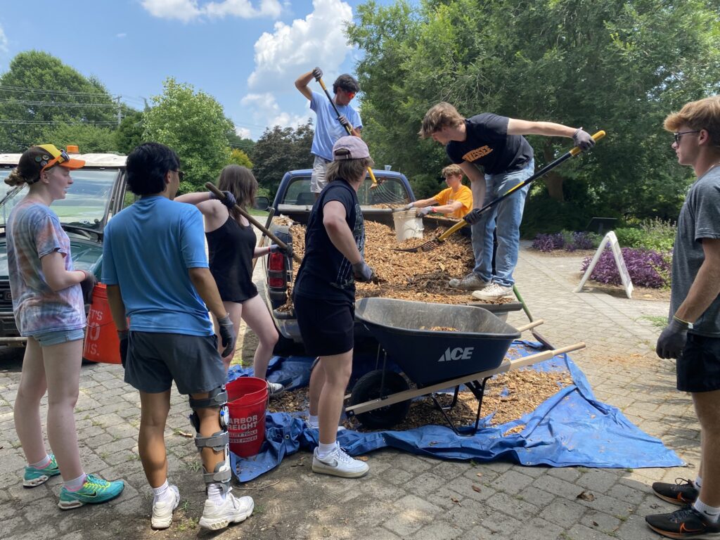 a group of volunteers empties out a truck load of mulch into a wheelbarrow to apply to an established flower bed